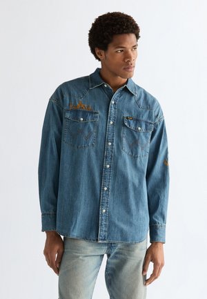 Ung mann iført en blå denimskjorte med knapper og broderte brystlommer, som ser litt mot høyre mot en enkel lys bakgrunn.