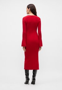 Rød ribbestrikket kjole med lange, utsvingte ermer, tettsittende silhuett, som når midt på leggen, kombinert med høye svarte støvletter.