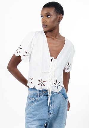 Junge Frau in weißer kurzärmliger Bluse mit floralen Ausschnitten und Knoten vorne, kombiniert mit hellblauen, hoch taillierten Jeans, schaut zur Seite.