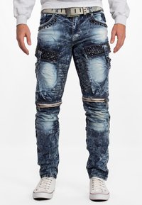 Pantalons cargo en denim bleu clair délavé avec plusieurs poches, détails en zip et rivets métalliques le long de la taille et des poches.