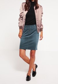Chaqueta bomber de satén en rosa claro, combinada con una falda lápiz en verde azulado con botones frontales. La modelo lleva zapatos negros sin cordones.