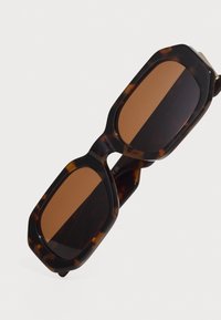 Lunettes de soleil en écaille de tortue avec des montures octogonales surdimensionnées et des verres teintés marron. Texture lisse avec un motif subtil sur la monture.