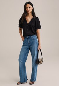 Črna bluzica s kratkimi napihnjenimi rokavi in gumbi, v kombinaciji z modrimi širokimi hlačami z visokim pasom. Dodatki vključujejo majhno črno in kremno torbico.