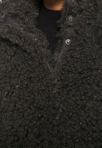 Manteau noir à texture, fabriqué en matériau pelucheux avec une fermeture éclair et des boutons-pression sur le devant. Il dispose d'un col haut et d'un intérieur en tricot côtelé.