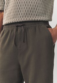 Dark olive green pants featuring an elastic waistband with a drawstring, textured fabric, and side pockets. Patterned top not fully visible.