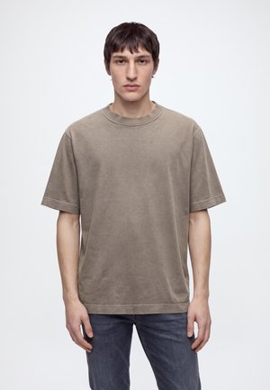 Un homme porte un tee-shirt ample à manches courtes couleur taupe et un jean foncé sur un fond clair uni.