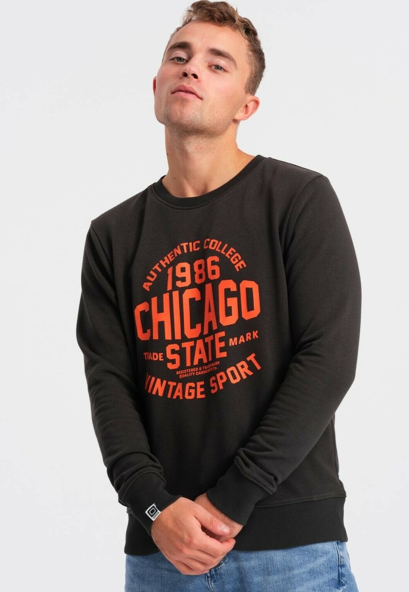 Maglione nero con maniche lunghe e scollo a girocollo. Presenta una grafica in stile vintage rossa con la scritta "Chicago State 1986 Vintage Sport."