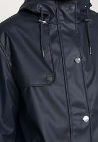 Marinblå vattentät jacka med huva, utrustad med silverfärgad hårdvara, tryckknappar och justerbara dragskor. Slät, glänsande textur.