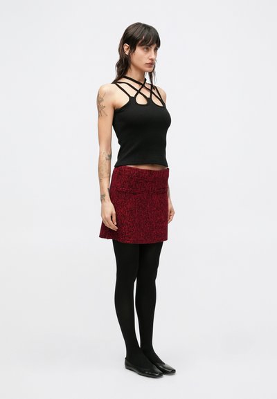 Zwart geribd tanktop met gekruiste bandjes, gecombineerd met een rode textuurrijke minirok. Model draagt zwarte panty's en platte schoenen.