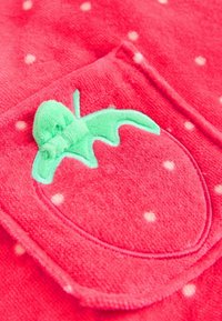 Rdeče flis tkanine z belimi pikami, z zelenim izvezenim vzorcem jagode z mašnjo na žepu. Mehka tekstura in igriv vzorec.