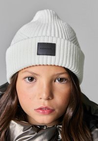 Vit ribbad stickad mössa med en svart läderlapp med präglad text. Designen är tajt och har en något spetsig topp.