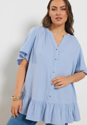 Frau trägt eine hellblaue, geknöpfte Kurzarmbluse mit Rüschensaum, kombiniert mit blauen Jeans und goldenen Armreifen am Handgelenk.