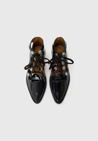 Schwarze Lackleder-Stiefeletten mit spitzen Zehen, schnürbarem Vorderteil, dekorativen silbernen Beschlägen und kontrastierendem, braunem Innenraum.