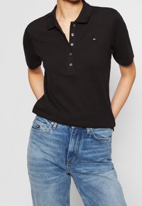 Polo noir avec une patte de boutonnage à trois boutons, fabriqué en tissu coton, associé à un jean en denim bleu clair avec un petit logo sur la poche.