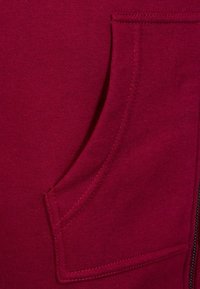 Tejido de sudadera burdeos con un bolsillo lateral con costura plana y una textura suave; detalle de cremallera visible en el lado derecho.