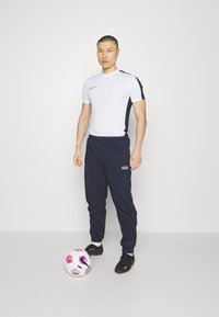 Weißes Sporthemd mit kurzen Ärmeln und schwarzen Akzenten, kombiniert mit marineblauen Hosen. Schwarze Fußballschuhe und ein mehrfarbiger Fußball sind vorhanden.
