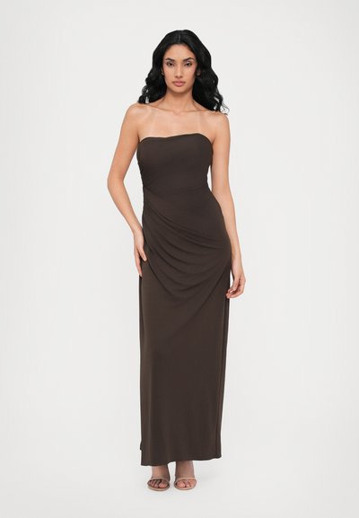 Abito maxi marrone senza spalline con tessuto drappeggiato, texture liscia e design aderente. La modella indossa sandali aperti.