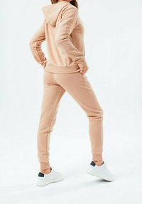 Světle růžová mikina s kapucí a odpovídající tepláky vyrobené z měkké tkaniny, s žebrovanými manžetami a stahovací šňůrkou, doplněné o bílé tenisky.