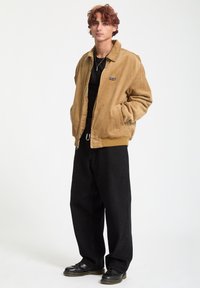 Veste en velours côtelé brun clair avec un col pointu, fermeture éclair à l'avant, poches latérales et un petit logo noir, associée à un pantalon large noir et des chaussures noires.