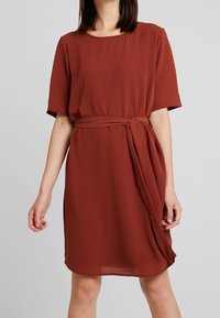 Une robe courte de couleur rouille, à manches courtes, avec un col rond, une taille ceinturée et un détail drapé à l'avant. Fabriquée à partir d'un tissu léger et texturé.