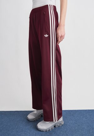 Oseba, ki nosi bordo Adidas trenirko s belimi črtami in logotipom, skupaj s sivimi mrežastimi supergami, stoji na modrem preprogi.