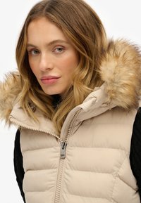 Beige gewatteerde vest met een ritsluiting en een met bont afgeronde capuchon. De stof heeft een zachte textuur met horizontale quiltsecties.