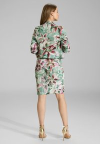 Ensemble veste et jupe à imprimé floral en vert pastel avec des fleurs roses et blanches. Présente un design structuré, une coupe ajustée et des talons dorés brillants.