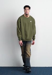 Olivno zelena pulover s belim Nike logotipom, v kombinaciji s pošvitanimi modrimi kavbojkami. Obleka ima sproščen kroj in vidne obrabe.