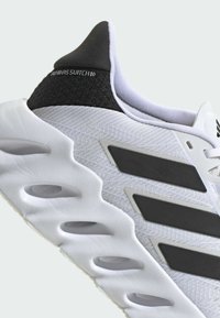 Scarpa sportiva bianca con tomaia texturizzata, applicazione nera sul tallone e tre strisce nere angolari. Presenta una suola spessa e imbottita con aperture.