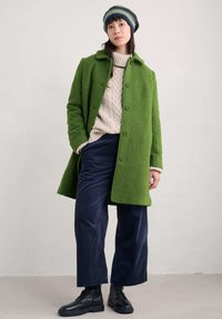 Grön texturerad ullkappa med knappstängningar, krämfärgad kabelstickad tröja, marinblå kordflöjdbyxor och svarta snörstövlar.