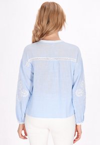 Blusa celeste chiaro con maniche lunghe, tessuto leggero, dettaglio in pizzo lungo le spalle e ricamo floreale bianco sulle maniche.