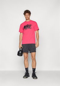 Lyserød Nike t-shirt med sort logo design, parret med mørkegrå shorts. Modellen holder en sort kettlebell og har sorte sneakers og sokker på.