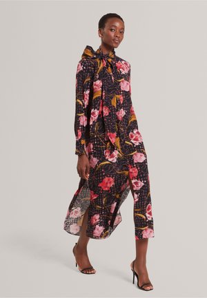 Kvinde iført en lang sort blomsterkjole med pink og røde blomster, sideslids, og sorte sandaler med høje hæle mod en ensfarvet baggrund.