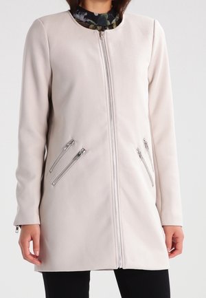 Vrouw draagt een lichtbeige getextureerde lange jas met rits aan de voorkant en vier diagonale ritssluitingen over een zwarte broek en een bloemen top.