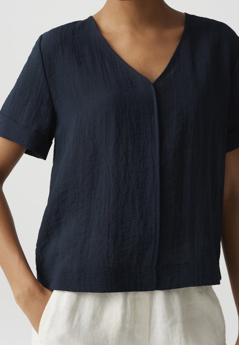 Femme portant un chemisier texturé bleu marine foncé à manches courtes avec col en V et pantalon blanc, debout avec une main dans la poche devant un fond uni.