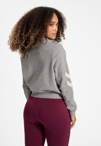Grauer Pullover mit weißen Streifen an den Ärmeln, gerippten Bündchen und einem lässigen Schnitt. Kombiniert mit burgunderfarbenen Leggings, vor einem weißen Hintergrund.