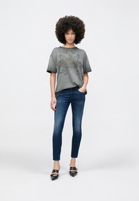 Camiseta gris de gran tamaño con gráfica de águila, combinada con jeans azules ajustados. Modelo lleva zapatos planos negros puntiagudos. Fondo blanco liso.