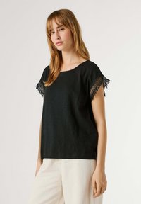 Kobieta o prostych, jasnobrązowych włosach, ubrana w czarną bluzkę z krótkim rękawem z koronkowym wykończeniem oraz kremowe spodnie, stojąca na jednolitym tle.