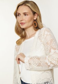 Cardigan bianco in pizzo aperto con motivi floreali, maniche lunghe e dettagli testurizzati, indossato sopra una maglietta bianca. Orecchini rotondi in tonalità terra.