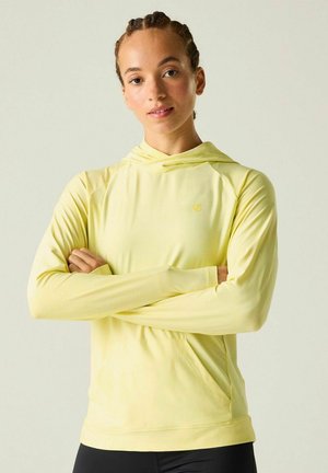 Woman with braided hair wearing a light yellow long-sleeve hoodie, arms crossed, standing against a plain light background.