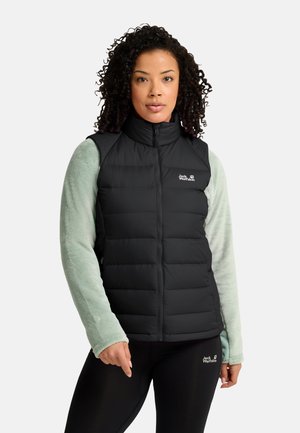 Frau trägt eine schwarze gesteppte Weste über einem hellgrünen Fleecepullover und schwarzen Leggings, steht mit neutralem Gesichtsausdruck vor einem einfarbigen Hintergrund.