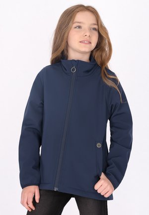 Junges Mädchen mit langen Haaren, trägt eine marineblaue Reißverschlussjacke, steht mit einer Hand in der Tasche und schaut leicht zur Seite.