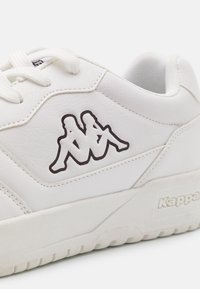 Weiße Ledersneaker mit strukturiertem Finish, ausgestattet mit einem schwarzen Grafiklogo von zwei sitzenden Figuren an der Seite. Schnürdesign, dicke Sohle.
