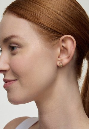 Złote kolczyki w kształcie obręczy z małym akcentem w postaci liścia, noszone przez osobę o czerwonych włosach, podkreślające wzór i blask kolczyków.