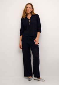 Completo due pezzi in pizzo blu navy composto da una maglietta cropped con bottoni e maniche ¾, abbinata a pantaloni a gamba larga. Accentuato con scarpe slip-on argentate.