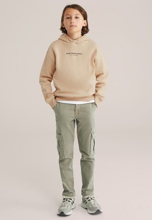 Kind staat rechtop met het gezicht naar voren, gekleed in een beige hoodie, groene cargobroek en witte sneakers, met handen in de zakken, tegen een effen achtergrond.