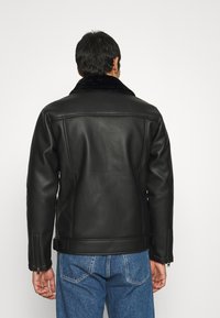 Giubbotto di pelle nera con colletto in pelliccia, polsini con zip e taglio dritto, abbinato a jeans azzurri chiari e una finitura liscia.