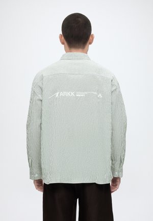 Άνδρας που φαίνεται από πίσω, φοράει ένα μακρυμάνικο πουκάμισο ανοιχτού γκρι και λευκού ριγέ με τη λέξη «ARKK» και γραφικό βέλος στην πλάτη.