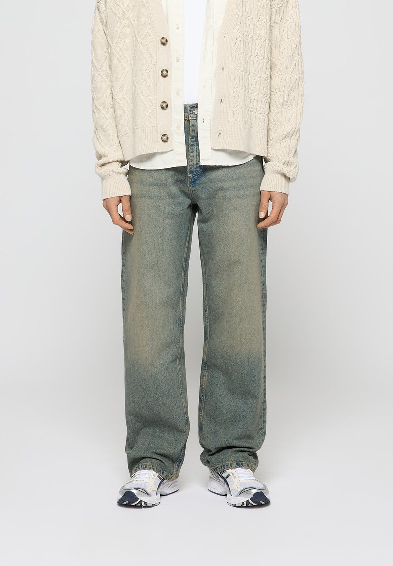 Beige kabelstrikket cardigan over en hvid skjorte, båret med lyseblå lige ben jeans og hvide sko med marineblå detaljer.