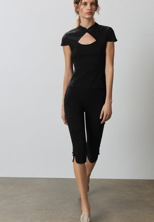 Femme marchant portant un haut noir ajusté avec un col goutte et un pantalon noir raccourci avec des boutons décoratifs à l'ourlet.
